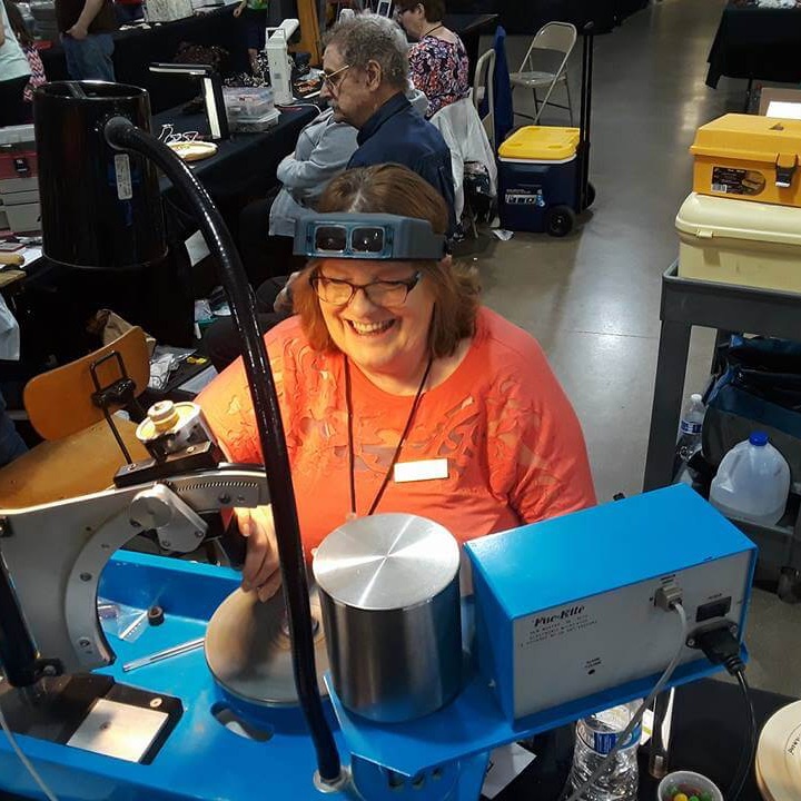 Smiling woman wearing magnifying lenses sits behind a blue faceting machine at a gem show.
