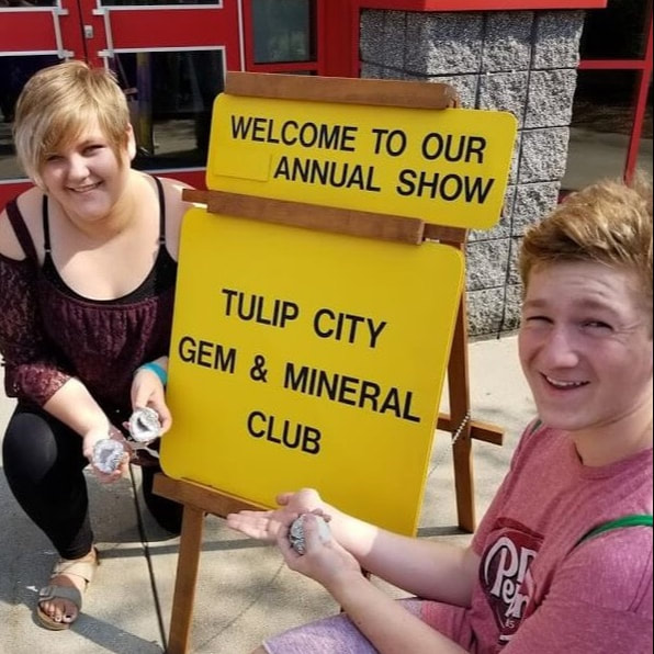 Two smiling teens holding cracked geodes crouch next to a yellow sign with the words 